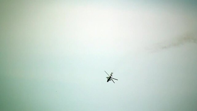 A single helicopter ascends into the pale blue sky, leaving a trail of smoke or cloud behind it. The sky is mostly clear with light hazy clouds, emphasizing the helicopter's solitary flight.