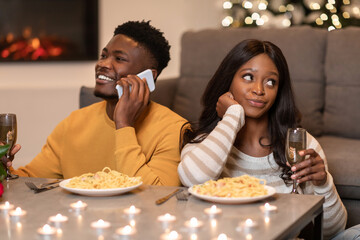 A woman sits at a dining table, looking bored while her partner is engaged on a cellphone during...