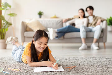 A young girl lies on her stomach with a piece of paper, coloring with crayons. Her parents sit on a...