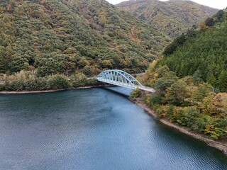 A bridge spans a river with a blue color. The bridge is surrounded by trees and mountains. The water is calm and clear
