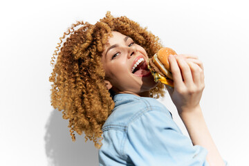 Young woman with curly hair enjoying a burger, showcasing joy and indulgence with a vibrant smile...