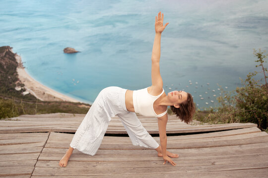 Young woman practicing Revolved Triangle Pose on wooden platform by ocean. Fit lady maintains balance and stretching gracefully surrounded by nature and serenity