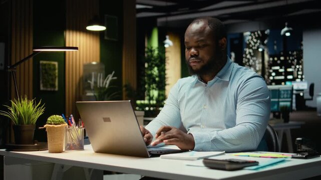 African american guy typing information on laptop and checking reports, proofreading and reviewing data for a new business objective or target. Company management after hours. Camera A.