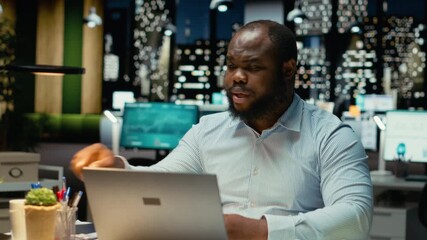 Black businessman engaging in online video call from dark office, maintaining eye contact on webcam. Attending meeting for meaningful conversation, building rapport with investors. Camera B.
