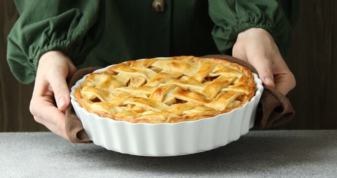 Woman putting baking dish of delicious homemade apple pie on grey textured table against dark wooden background, closeup