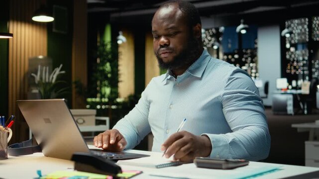 African american employee drafting updates and handwriting a summary late at night at the office. Data reports information with the monthly activity planner, business goals and agenda. Camera A.