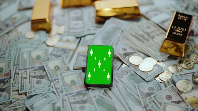 Smartphone with a chroma key green screen displaying candlestick charts for trading, resting on a pile of one-hundred-dollar bills, gold bars, and coins, representing wealth and finance