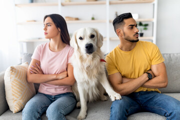 A man and woman sit on a couch, looking away from each other, while their golden retriever sits...