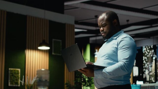 African american guy proofreading a document before transcribing on a textbook, extracting the main ideas from reports on laptop. Expert documenting after hours late at night. Camera B.