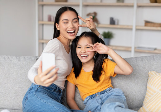 A woman and a young girl share smiles as they take selfies on a soft sofa. The bright living room has shelves and a cheerful atmosphere, showcasing their close bond and happiness. - Powered by Adobe