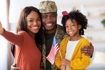 A joyful family gathers in a warm living room, celebrating a military homecoming. The father in uniform stands with his wife and daughter, all smiling and holding a flag.