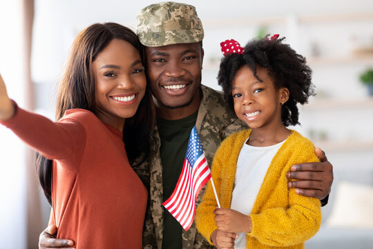 A joyful family gathers in a warm living room, celebrating a military homecoming. The father in uniform stands with his wife and daughter, all smiling and holding a flag. - Powered by Adobe