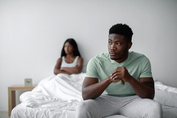 Obraz premium A man sits on the edge of a bed looking pensive while a woman in the background sits with her arms crossed, indicating emotional distance in a quiet setting.