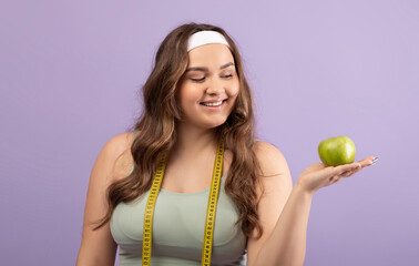 A cheerful millennial European plus size female in sportswear smiles while holding a green apple in one hand.