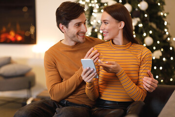 A young couple sits close together on a cozy couch, smiling and looking at a phone. The warm glow of a fireplace and Christmas tree lights create a festive atmosphere.