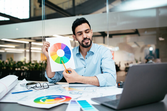 A man in a blue shirt holds a color wheel while sitting at a table covered with design plans and documents. He appears focused and engaged in the discussion.