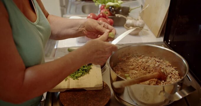 Captivating close-up of a woman's hands skillfully chopping fresh okra, incorporating the vibrant green into a rich, simmering pot of ground meat on the stove.
