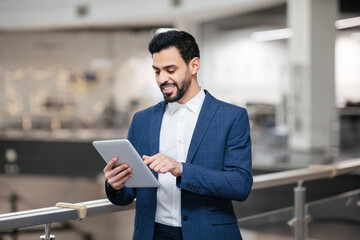 Fototapeta premium A man in a suit engages with a tablet, smiling as he stands in a contemporary office space with bright lighting and minimalistic design.