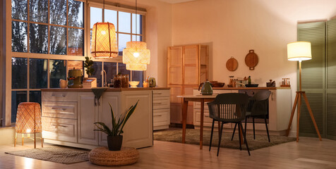 Interior of modern kitchen with counters, dining table and glowing lamps in evening