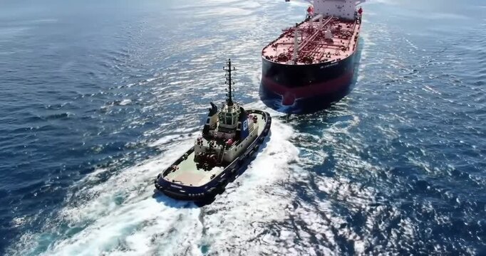 Aerial view of a powerful tugboat maneuvering a large red cargo ship in the open blue sea.