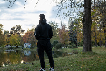 man in black hoodie and cargo pants standing by a tranquil lake in an autumn park, contemplating the scenic nature and colorful trees at sunset