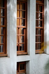 Authentic architectural detail of three parallel brown wooden multi-pane windows with small square window on gray concrete wall, natural sunlight casting shadows and illuminating window frames.