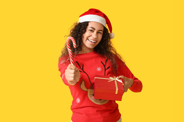 Happy young African-American woman in Christmas sweater with candy cane and gift box on yellow background