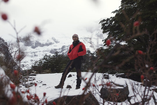 A man stands confidently in a snow-covered mountain setting, framed by trees and distant peaks. His red jacket contrasts with the white snow, symbolizing energy and adventure in the middle of winter e