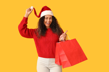 Beautiful young African-American woman in Santa hat with shopping bags and candy cane on yellow background. Christmas celebration