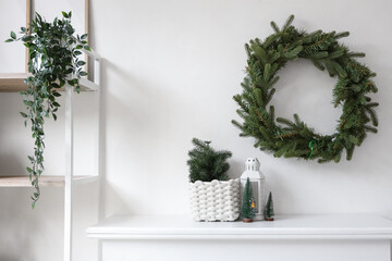 Mantelpiece with decorative Christmas trees and glowing lantern near white wall