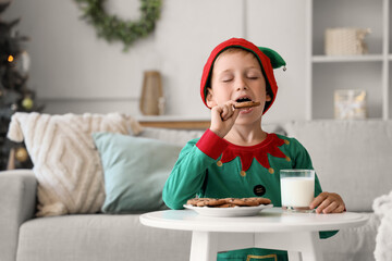 Cute little boy dressed as Christmas elf eating cookies with cup of milk at home