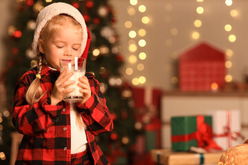 Cute little girl in Santa hat drinking milk at home on Christmas eve