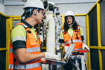 Engineers in safety gear collaborating, using a laptop to troubleshoot or program a robotic arm in a factory. Industrial technology, and the integration of software into modern manufacturing process