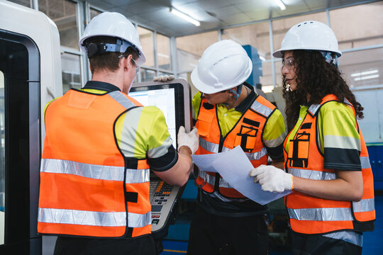 Team of three industrial engineers in safety gear are collaborating to operate a CNC machine, reviewing technical documents and discussing production data on the factory manufacturing