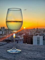 A glass of white wine rests on a stone railing overlooking a breathtaking view of Buenos Aires at sunset. The golden light illuminates the cityscape, creating a warm and elegant atmosphere.