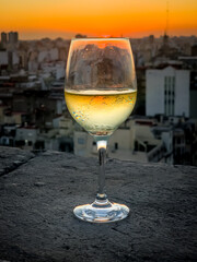 A glass of white wine rests on a stone railing overlooking a breathtaking view of Buenos Aires at sunset. The golden light illuminates the cityscape, creating a warm and elegant atmosphere.