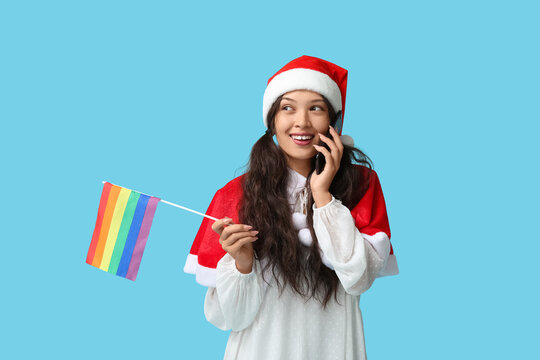 Young Asian woman in Santa hat with LGBT flag talking by mobile phone on blue background - Powered by Adobe