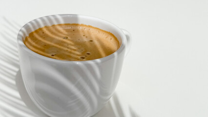 A cup of fresh coffee on a white table and shaded by plants.