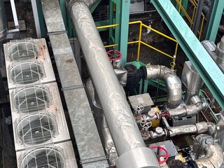 Aerial view of a commercial rooftop mechanical area, featuring large cylindrical insulated HVAC piping, multiple cooling condenser units, complex valve arrangements, and yellow safety railings 