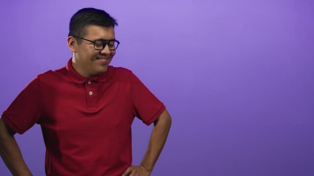 Hispanic man wearing glasses and a red polo, smiling with hands on hips in a purple studio; confidence.