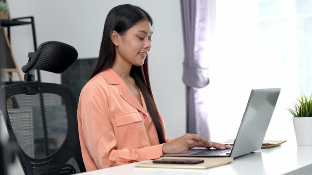 Asian young woman working laptop home desk wearing peach blouse with notebook and smartphone nearby smiling while typing bright modern office ergonomic chair beside potted plant natural light focused