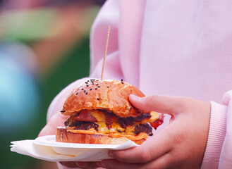 A young woman carrying a gourmet burger with melted cheese on a tray with napkins, a close-up image at Karlin food festival in Prague.