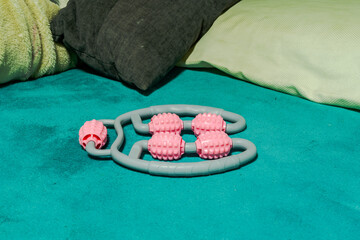 Handheld massage roller with pink textured wheels on teal bed surrounded by pillows, showcasing...
