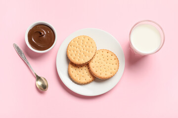Tasty sandwich biscuits with chocolate cream and glass of milk on pink background