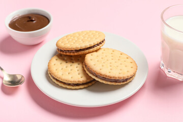 Tasty sandwich biscuits with chocolate cream and glass of milk on pink background