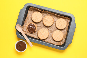 Baking dish with tasty sandwich biscuits and bowl of chocolate cream on yellow background