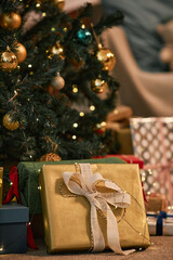 Christmas tree decorated with gold ornaments standing beside wrapped gifts in various sizes and colors, golden present with ribbon placed in foreground, festive holiday setting