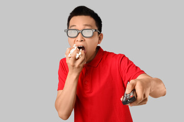 Emotional young Asian man with remote controller and popcorn watching TV on grey background