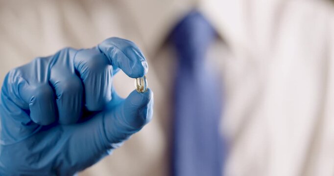  a hand wearing a blue medical exam glove, holding a golden, translucent gel capsule or supplement. The reflective surface of the pill shows a miniature, reflection of the person as the camera tracks