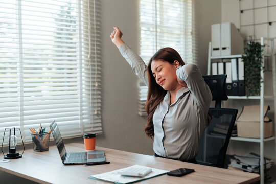 Asian woman feeling neck pain and fatigue, stretching at her office desk from long hours of work and computer use